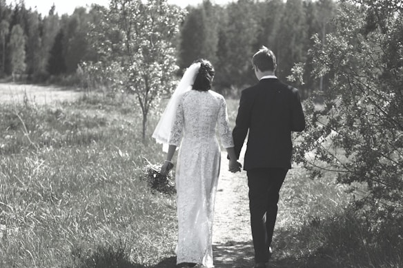 A couple is walking hand in hand along a grassy path, surrounded by lush greenery and trees. The woman is wearing a white gown with a veil, holding a bouquet, and the man is in a dark suit. The setting appears serene and natural, with a soft, muted color palette.