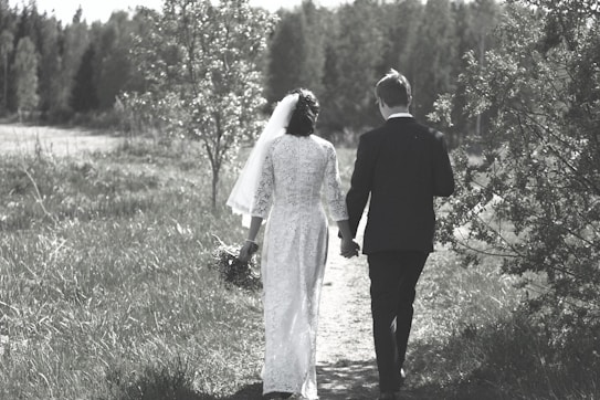 A couple is walking hand in hand along a grassy path, surrounded by lush greenery and trees. The woman is wearing a white gown with a veil, holding a bouquet, and the man is in a dark suit. The setting appears serene and natural, with a soft, muted color palette.