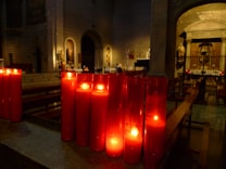 A dimly lit church interior with a row of red votive candles glowing softly in a darkened atmosphere. Architectural details like arches, stone walls, and religious icons can be seen in the background, contributing to a serene and solemn ambiance.