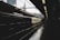 A commuter train is situated on a dimly lit urban platform during the day. The train's exterior is illuminated by overhead lighting, contrasting with the shadows in the station. Skyscrapers are visible in the distant background, contributing to the industrial cityscape.