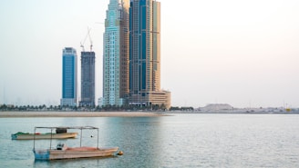 High-rise buildings stand tall along a calm waterfront, with a couple of small boats anchored in the foreground. The sky is clear and slightly overcast, giving a serene and peaceful atmosphere. Construction cranes are visible on some of the buildings, indicating ongoing development.