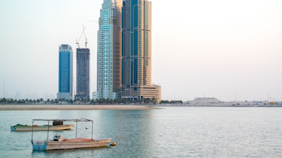 High-rise buildings stand tall along a calm waterfront, with a couple of small boats anchored in the foreground. The sky is clear and slightly overcast, giving a serene and peaceful atmosphere. Construction cranes are visible on some of the buildings, indicating ongoing development.