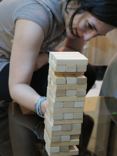 A person is intensely focused on a tower made of wooden blocks, possibly playing a game of Jenga. The person has dark hair and is leaning forward with a bracelet on their wrist, carefully considering their next move.