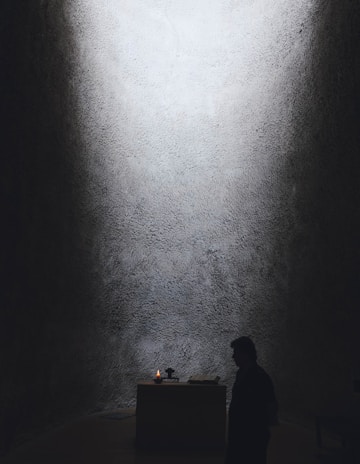 A dimly lit space with textured walls and a small altar at the center. A candle is flickering on the altar, providing the primary source of light. A person stands in silhouette nearby, adding to the solemn and contemplative atmosphere.