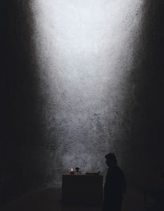 A dimly lit space with textured walls and a small altar at the center. A candle is flickering on the altar, providing the primary source of light. A person stands in silhouette nearby, adding to the solemn and contemplative atmosphere.
