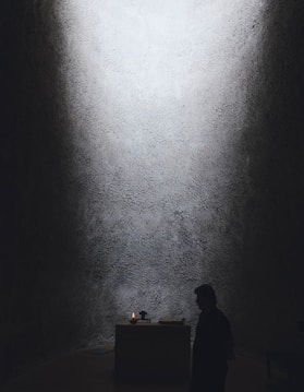 A dimly lit space with textured walls and a small altar at the center. A candle is flickering on the altar, providing the primary source of light. A person stands in silhouette nearby, adding to the solemn and contemplative atmosphere.