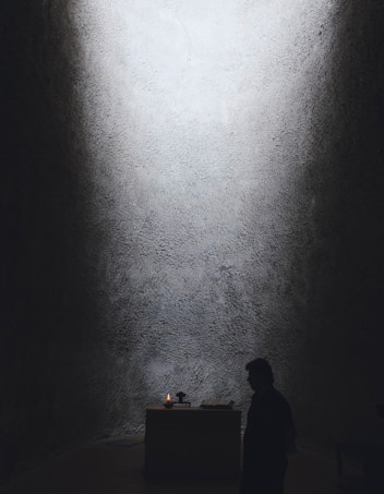 A dimly lit space with textured walls and a small altar at the center. A candle is flickering on the altar, providing the primary source of light. A person stands in silhouette nearby, adding to the solemn and contemplative atmosphere.