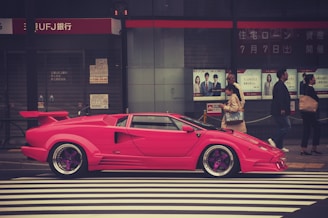 A bright pink sports car is parked on a street in front of a bank with Japanese signage. Several pedestrians are walking by, including a woman with a handbag and a man in a suit. The setting is urban with a crosswalk in the foreground.