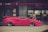 A bright pink sports car is parked on a street in front of a bank with Japanese signage. Several pedestrians are walking by, including a woman with a handbag and a man in a suit. The setting is urban with a crosswalk in the foreground.