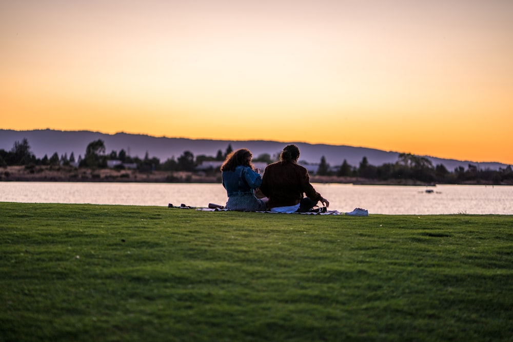 man and woman sitting on grass while fronting body of water