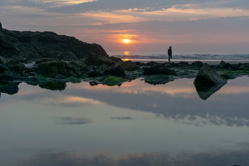 person walking beside body of water during sunset