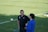 Two people are standing on a grassy soccer field, having a conversation. One person is wearing a black shirt with a logo, and the other is wearing a blue top. Soccer balls and training cones are scattered across the field.