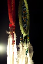 Two dreamcatchers hang against a dark background. The one on the left is red with white feathers and multicolored beads, while the one on the right is green with blue webbing and similar bead and feather decorations. A soft light highlights the textures and colors of the dreamcatchers.