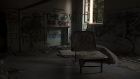 A dimly lit, abandoned room filled with debris and graffiti-covered walls. A broken chair is positioned near the center, and light streams through a partially open window, illuminating parts of the room. Shadows dominate, creating a sense of neglect and decay.