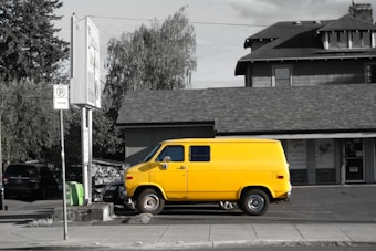 A bright yellow van is parked on a street in front of a gray building. The scene features a mix of commercial and residential architecture, with trees and signs around. The color of the van stands out vividly against the otherwise monochromatic setting.