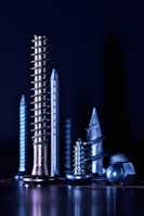 A group of various metallic screws and bolts, arranged vertically against a dark background, displaying different shapes and sizes with reflective surfaces.