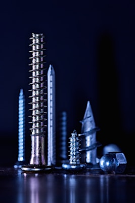 A group of various metallic screws and bolts, arranged vertically against a dark background, displaying different shapes and sizes with reflective surfaces.