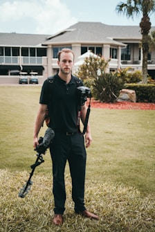 A person stands on a grassy lawn in front of a large building with a camera and stabilizer equipment. The structure in the background is a multi-story house surrounded by landscaped gardens and palm trees.