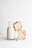 A glass bottle filled with milk is placed on a light-colored wooden surface. Next to the bottle, there are two slices of a crumbly cake with a reddish filling or topping, possibly raspberry. The background is plain and white, creating a minimalistic look.