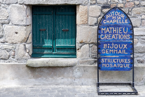 A rustic stone wall features a closed green wooden window with a weathered stone sill. To the right, there is a blue and black metal sign with French text, promoting artistic creations such as jewelry and mosaics.