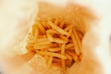 A top view of a paper bag filled with freshly cooked French fries. The fries appear golden and crispy, and the bag has a slightly crumpled texture.