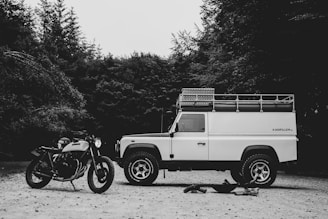 A black and white photograph depicts a vintage motorcycle parked next to a classic off-road vehicle. Both are situated on a gravel area surrounded by dense trees.