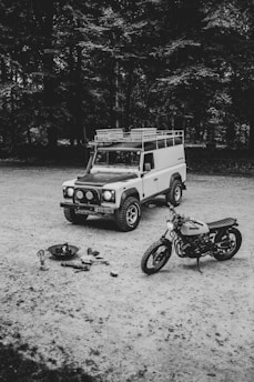 A vintage off-road vehicle and a motorcycle are parked on a gravel surface, with a small campfire setup including logs, a kettle, and cooking utensils nearby. The scene is set against a backdrop of dense trees, suggesting a peaceful and rustic outdoor environment.