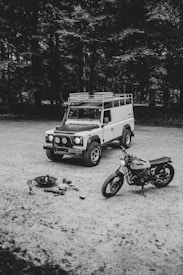 A vintage off-road vehicle and a motorcycle are parked on a gravel surface, with a small campfire setup including logs, a kettle, and cooking utensils nearby. The scene is set against a backdrop of dense trees, suggesting a peaceful and rustic outdoor environment.