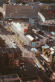 Aerial view of a cityscape featuring a large multi-story building with 'UQAM' written on it. The scene captures streets lined with trees and numerous people walking. There are buses and cars along the roads and smaller buildings with various architectural styles, including a modern complex with large glass facades.