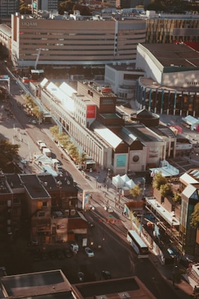 Aerial view of a cityscape featuring a large multi-story building with 'UQAM' written on it. The scene captures streets lined with trees and numerous people walking. There are buses and cars along the roads and smaller buildings with various architectural styles, including a modern complex with large glass facades.
