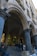 Stone arches and columns form a covered gallery, with people standing and interacting in a shaded area. Various framed artworks and boxes are displayed on the ground, suggesting a street market or outdoor exhibition. The architecture features classical design elements with detailed stonework.