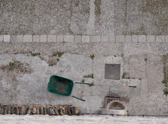 A wheelbarrow with a rusty green body lies on a textured cobblestone surface. Below the wheelbarrow, a neat stack of chopped firewood is arranged. A rectangular metal grate is embedded in the cobblestones nearby, with patches of grass peeking through some of the stone gaps.
