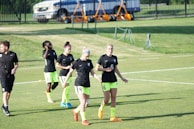 Several individuals wearing similar black shirts and neon green shorts are walking across a grassy sports field. Some are holding items, and they appear to be part of a sports team, possibly preparing or concluding practice. In the background, a fenced area and parked vehicles are visible.