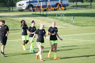 Several individuals wearing similar black shirts and neon green shorts are walking across a grassy sports field. Some are holding items, and they appear to be part of a sports team, possibly preparing or concluding practice. In the background, a fenced area and parked vehicles are visible.