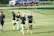 Several individuals wearing similar black shirts and neon green shorts are walking across a grassy sports field. Some are holding items, and they appear to be part of a sports team, possibly preparing or concluding practice. In the background, a fenced area and parked vehicles are visible.