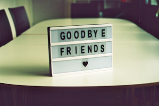 A lightbox sign with the message 'Goodbye Friends' and a heart symbol below is placed on an empty conference table. The room conveys a simple, calm atmosphere.