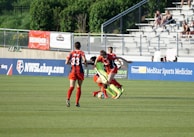 A soccer match scene with players in action on the field, one player attempting to control the ball while two opposing players challenge closely. The background includes a fence, advertising banners, and a small crowd sitting in bleachers.