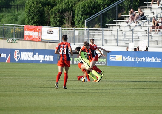 A soccer match scene with players in action on the field, one player attempting to control the ball while two opposing players challenge closely. The background includes a fence, advertising banners, and a small crowd sitting in bleachers.