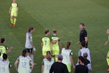 A group of female soccer players wearing bright green uniforms with black accents are gathered on a grassy field. Some players have numbers and names displayed on their jerseys. There is a referee or coach in black attire engaging with the team.