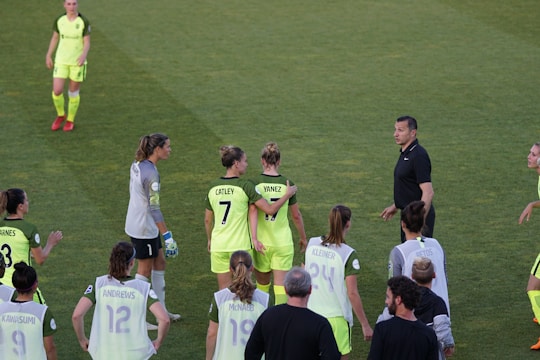 A group of female soccer players wearing bright green uniforms with black accents are gathered on a grassy field. Some players have numbers and names displayed on their jerseys. There is a referee or coach in black attire engaging with the team.