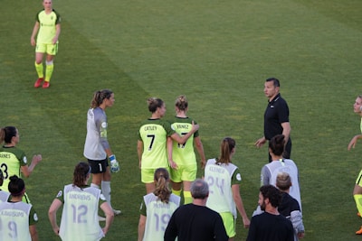A group of female soccer players wearing bright green uniforms with black accents are gathered on a grassy field. Some players have numbers and names displayed on their jerseys. There is a referee or coach in black attire engaging with the team.