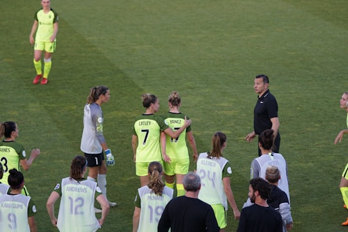 A group of female soccer players wearing bright green uniforms with black accents are gathered on a grassy field. Some players have numbers and names displayed on their jerseys. There is a referee or coach in black attire engaging with the team.