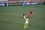 A soccer match taking place on a grassy field, featuring four players actively involved in the game. The players are wearing brightly colored jerseys, with one team in neon green and the other in red and blue. Advertising boards are visible in the background, indicating the location as Maryland SoccerPlex.