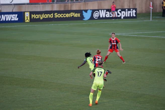 A soccer match taking place on a grassy field, featuring four players actively involved in the game. The players are wearing brightly colored jerseys, with one team in neon green and the other in red and blue. Advertising boards are visible in the background, indicating the location as Maryland SoccerPlex.