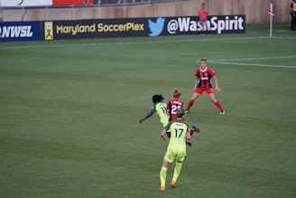 A soccer match taking place on a grassy field, featuring four players actively involved in the game. The players are wearing brightly colored jerseys, with one team in neon green and the other in red and blue. Advertising boards are visible in the background, indicating the location as Maryland SoccerPlex.