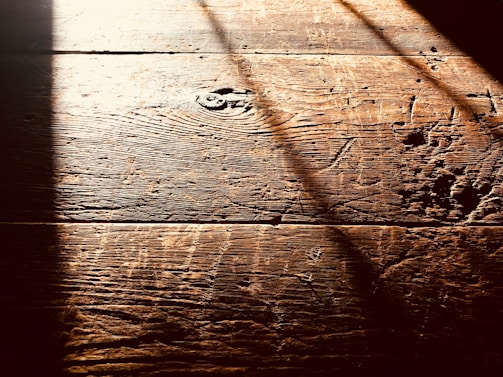 Warm, textured wooden floorboards with visible grain and shadows cast diagonally across the surface, creating a contrast between light and dark areas.