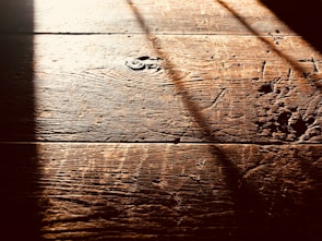 Warm, textured wooden floorboards with visible grain and shadows cast diagonally across the surface, creating a contrast between light and dark areas.