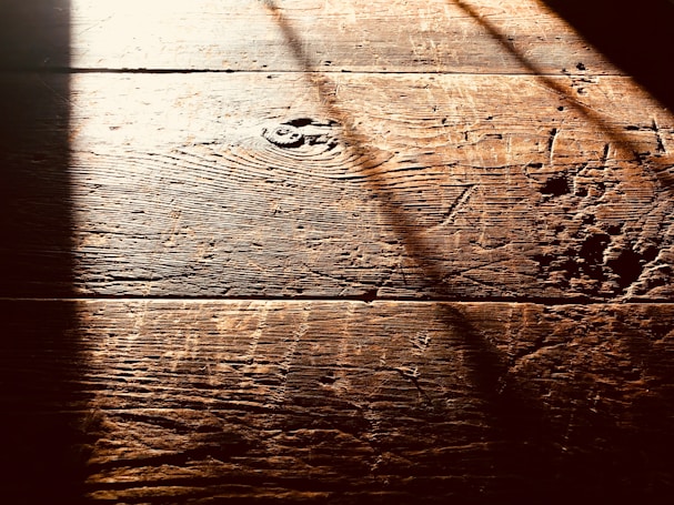 Warm, textured wooden floorboards with visible grain and shadows cast diagonally across the surface, creating a contrast between light and dark areas.