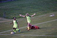 Two soccer players in bright green uniforms are on a grassy field. One player appears to be celebrating or signaling with raised arms, while another player runs with the ball close to the sideline. In the background, another player in a red uniform is lying on the ground. There are some equipment and gear placed near the edge of the field.
