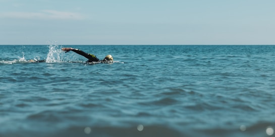 A person is swimming in a vast, open body of water with their head slightly above the surface, and their arm extended forward in mid-stroke. The water is calm with gentle waves, and the horizon line is visible in the distance under a clear sky.